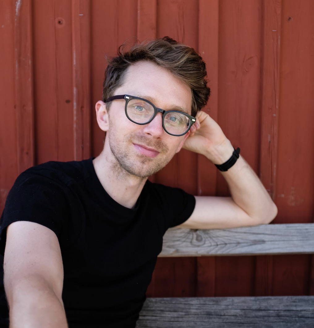 Portrait of Max Milne seated against a rust-red wall.